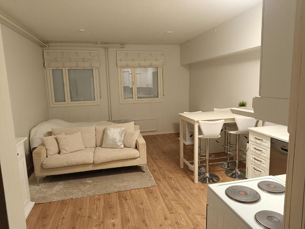 Huoneessa on sohva, ikkunat, ruokapöytä ja keittiö, valaistus on kirkas ja värit ovat vaaleita.